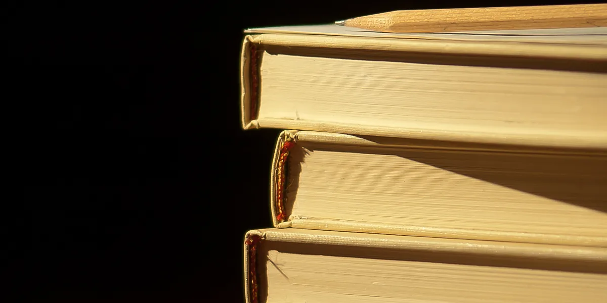 Tres libros de tapa dura apilados con un lápiz encima, iluminados desde un lado sobre un fondo negro
