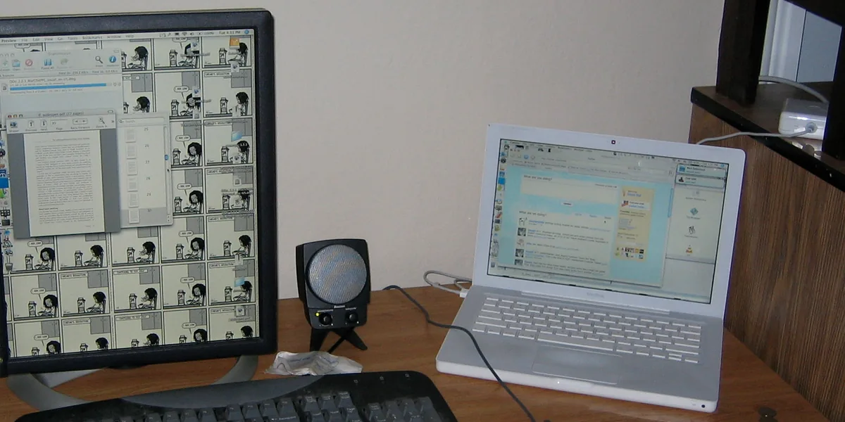 Monitor girado en vertical con un documento abierto, un altavoz pequeño, un teclado negro y un portátil blanco sobre una mesa de madera