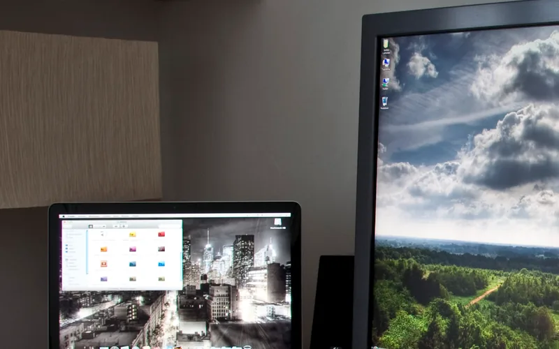 Pantalla de un portátil abierto a la izquierda de un monitor grande con un paisaje de nubes; detrás, una pared con un panel de tela