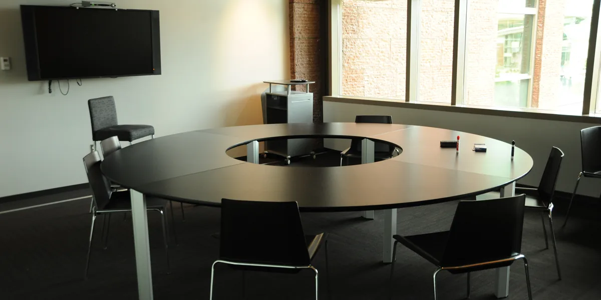 Sala con una pantalla negra colgada en la pared, una mesa redonda con sillas y un ventanal dividido en hojas a la derecha